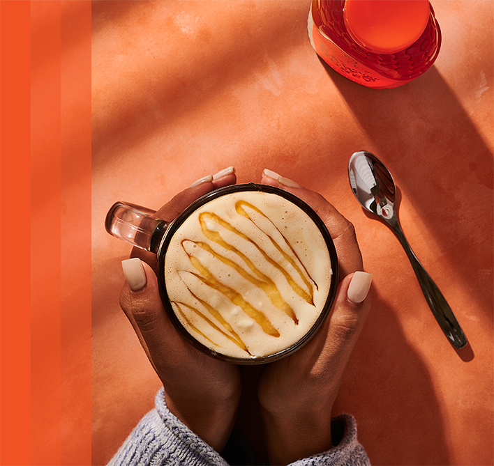 Hands holding clear mug of coffee with honey drizzle.