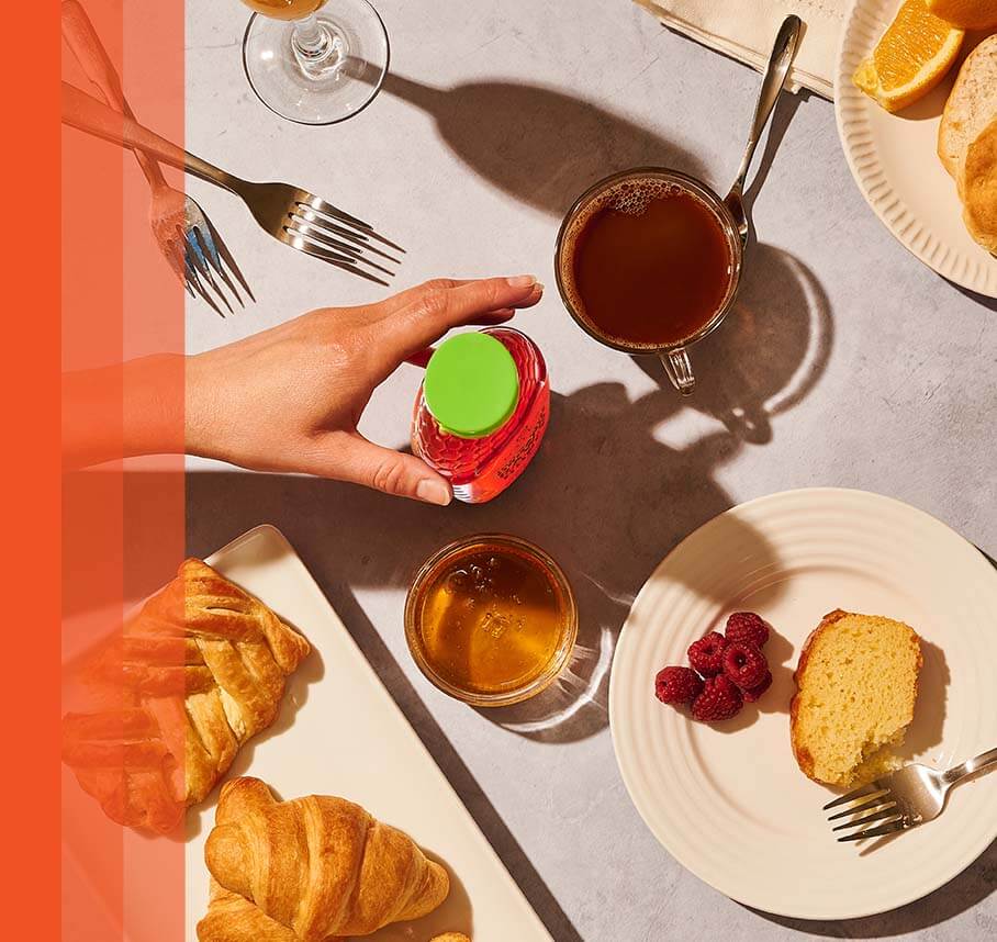 Hand reaching for a honey bottle on a breakfast table with pastries, honey, and fruit.