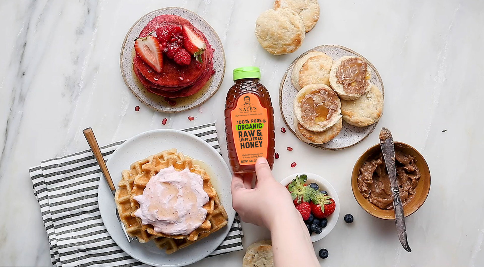 Hand holding a bottle of Nate's organic honey over waffles with cream, biscuits and berries on marble.
