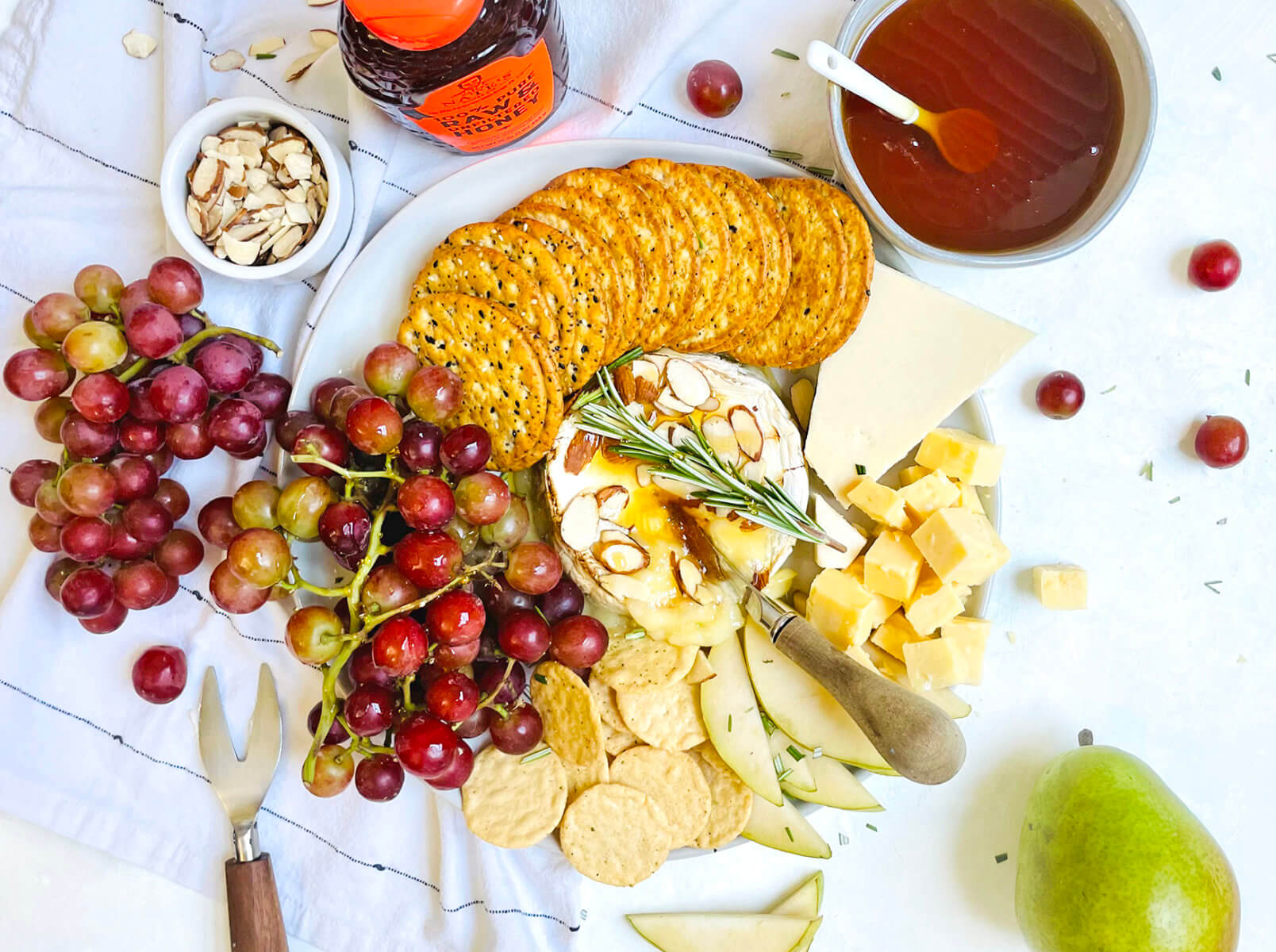 A cheese platter with grapes, crackers, nuts, honey, and a pear with a bottle of Nate's honey on a white cloth.