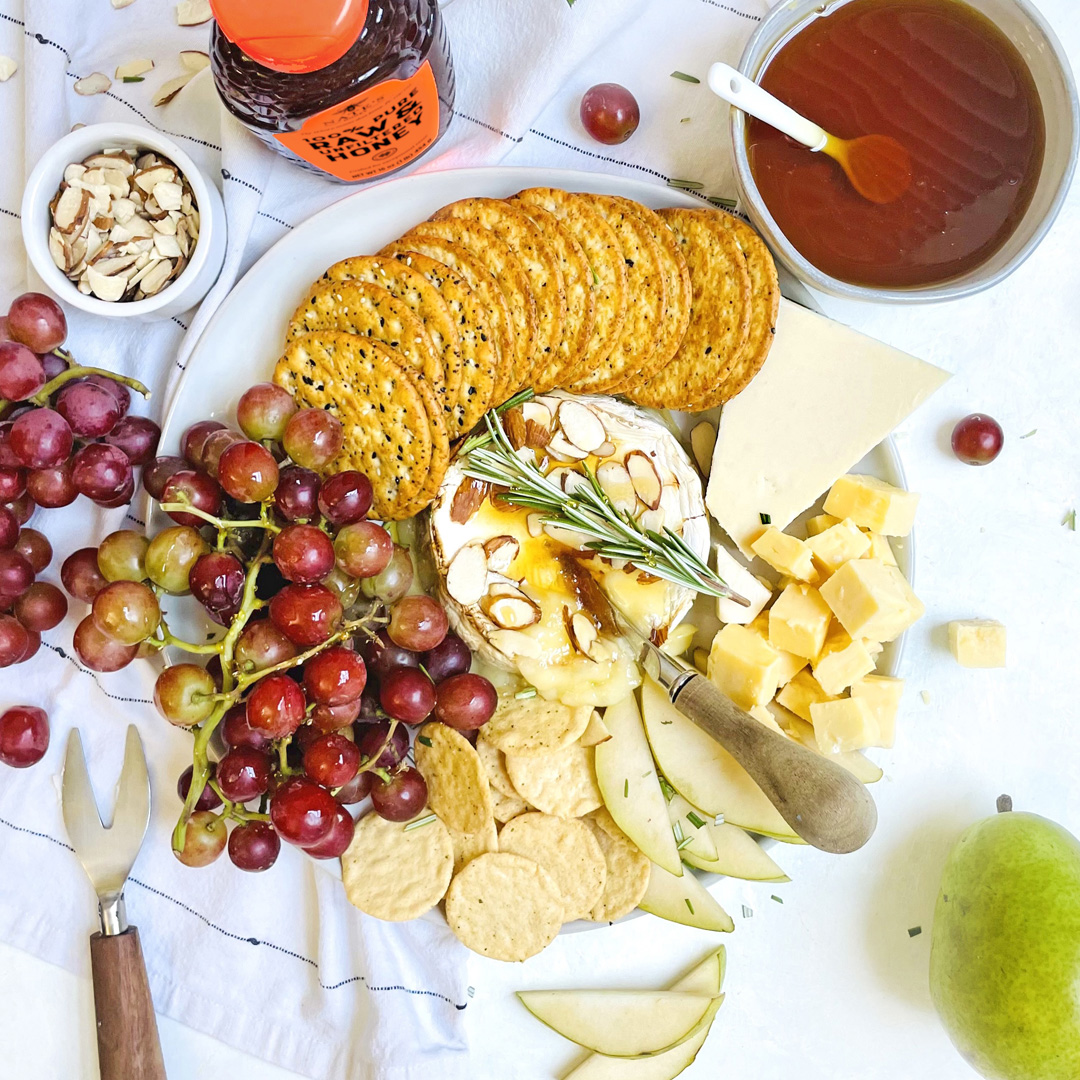 A cheese platter with grapes, crackers, nuts, honey, and a pear with a bottle of Nate's Honey on a white cloth.