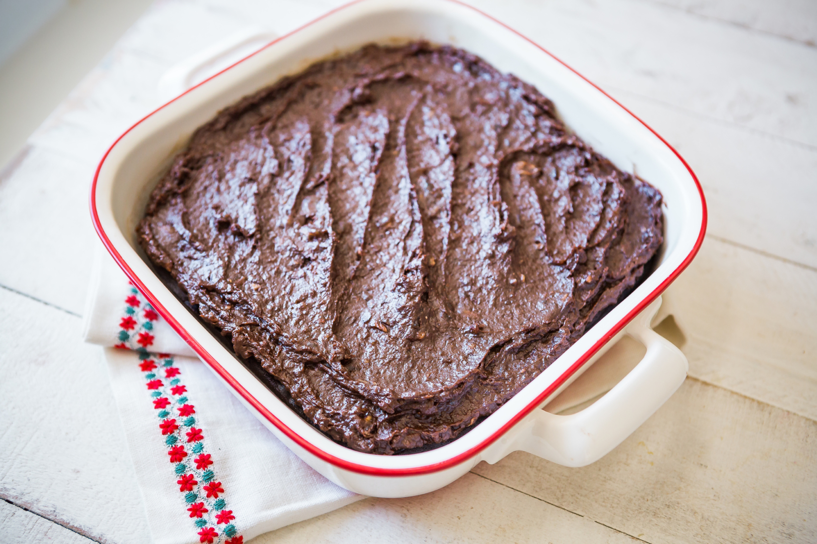 Freshly baked brownies in a white ceramic dish with red trim, on a white wooden table with a festive cloth.