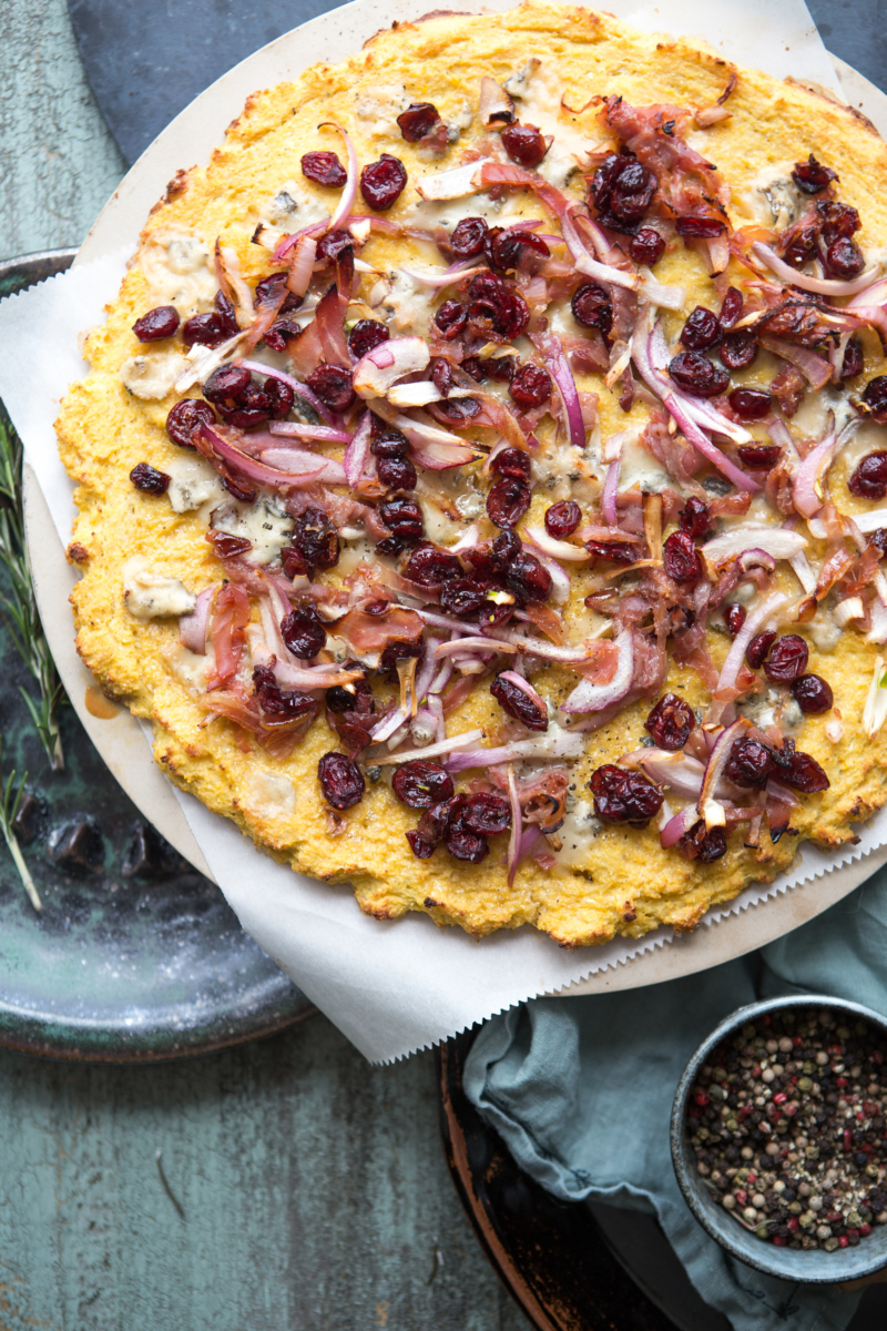 A rustic pizza topped with onions, prosciutto and cranberries and drizzled with honey, on a worn surface with pepper.