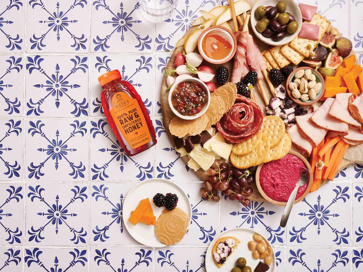 Charcuterie board with meats, cheeses, fruits, nuts and  a bottle of Nate’s honey on a patterned countertop.
