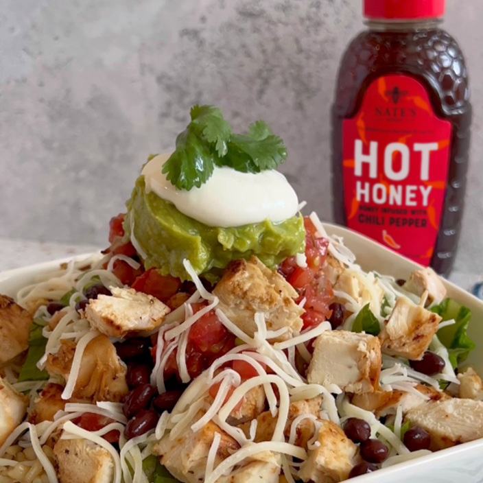 Cubes of grilled chicken in a bowl, topped with cheese, guacamole and sour cream, next to a bottle of Nate's Hot Honey.