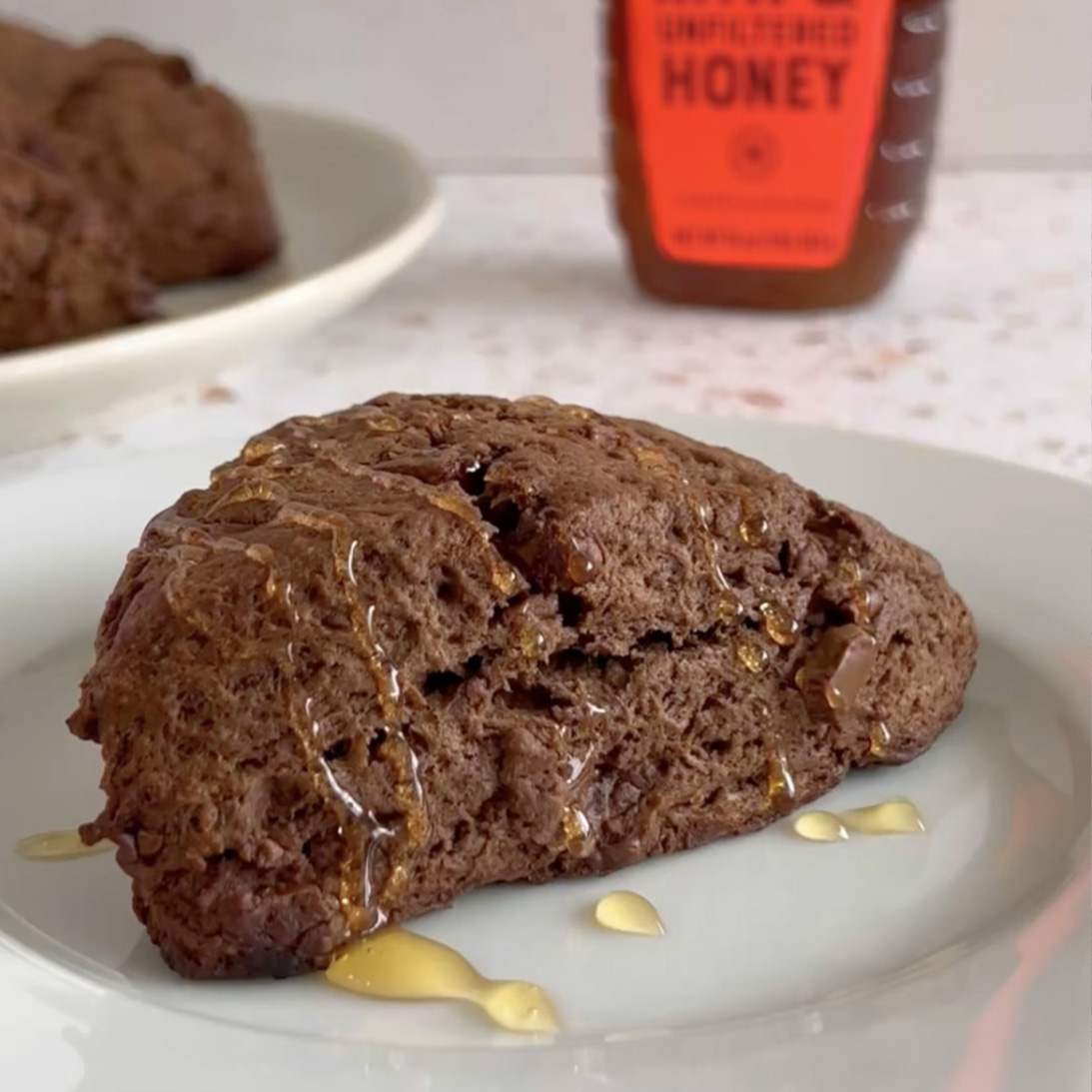 A chocolate scone drizzled with Nate's honey on a plate, with a bottle of Nate's honey in the background.