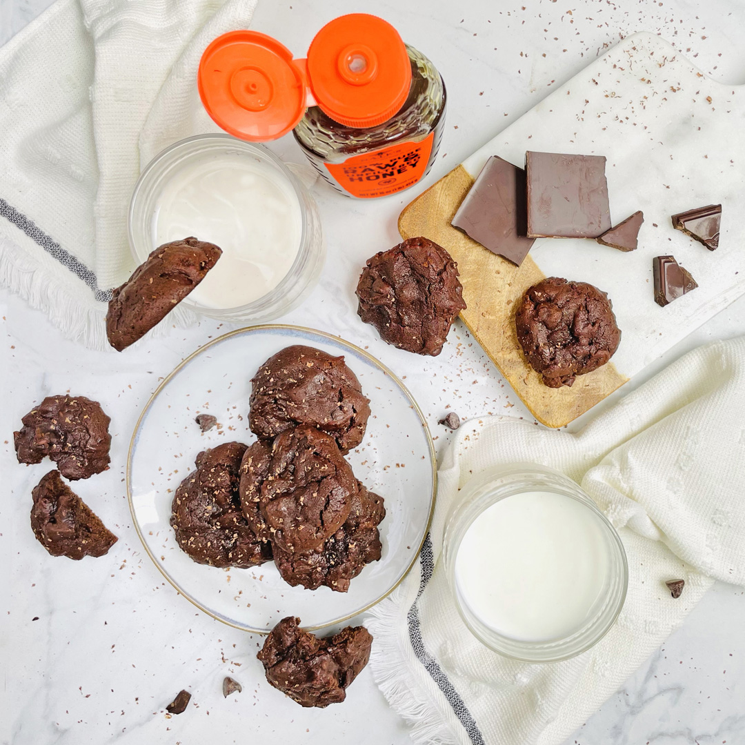 Chocolate cookies on a plate with chunks of chocolate, two glasses of milk, and a Nate’s honey bottle on marble.