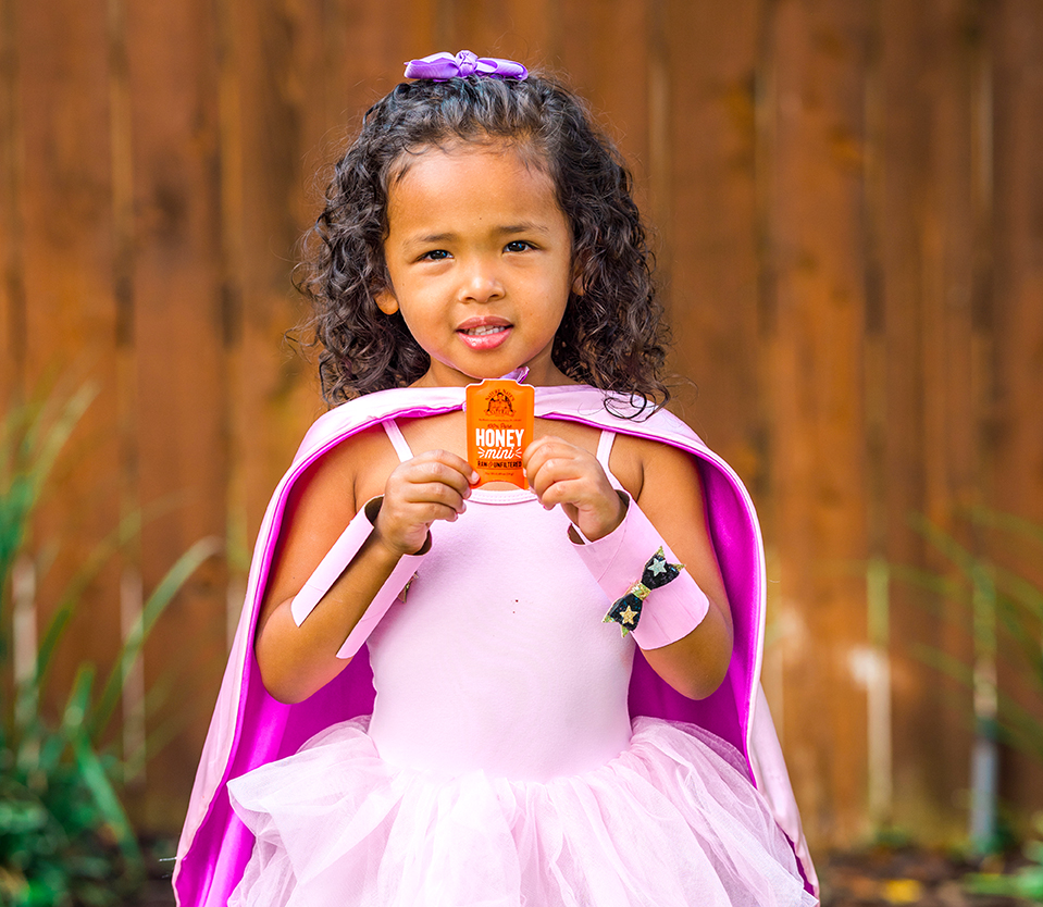 Young girl in a pink superhero costume holding a Nature Nate's honey packet, standing against a wooden fence.