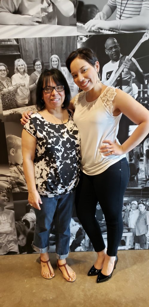 Two women posing for a photo together, one in a black and white top, the other in a white sleeveless top, both smiling.