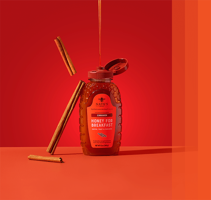 Bottle of Nate's Honey Cinnamon Honey for Breakfast with cinnamon sticks and a honey drizzle against a red background.
