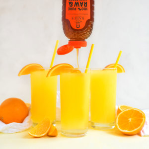Three glasses of citrus slushie with straws, garnished with orange slices, Nate's honey bottle pouring into one glass.