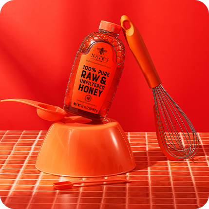 Nate’s Raw & Unfiltered Honey bottle with a whisk and honey spoon on a vibrant red background.