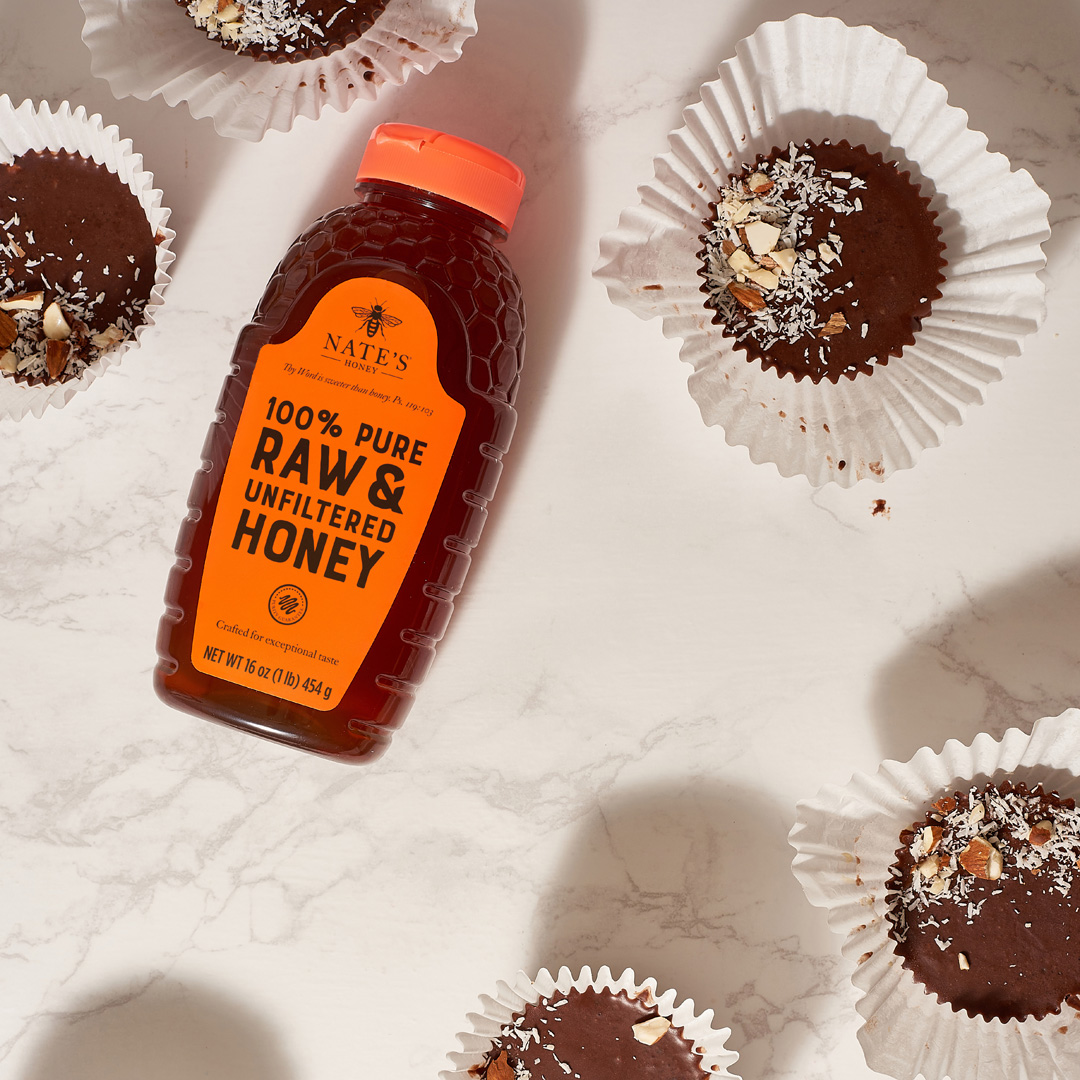 A bottle of Nate’s Honey surrounded by chocolate coconut almond treats on a marble surface.