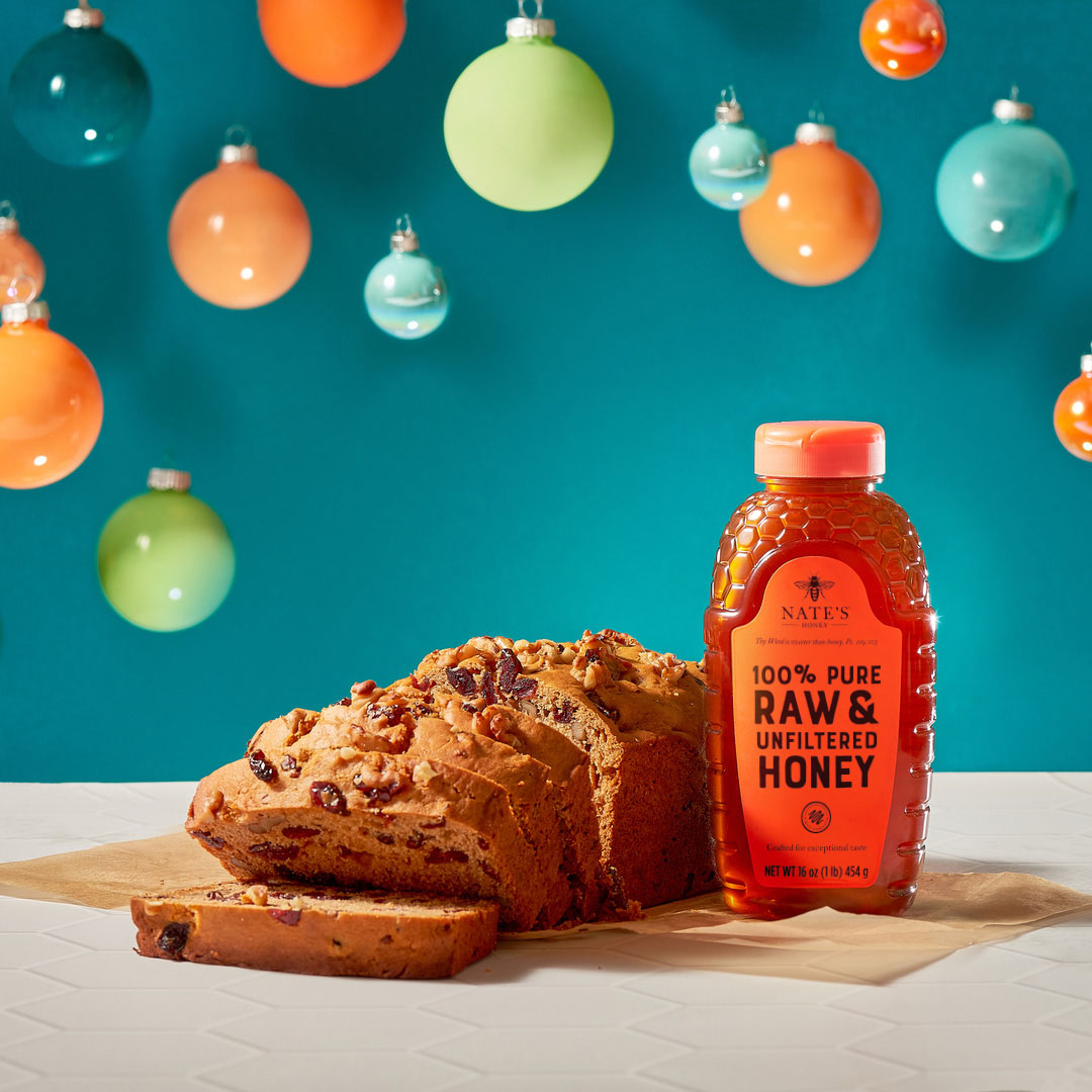 Cranberry nut bread beside a Nate’s Honey bottle, against a backdrop with colorful hanging ornaments.