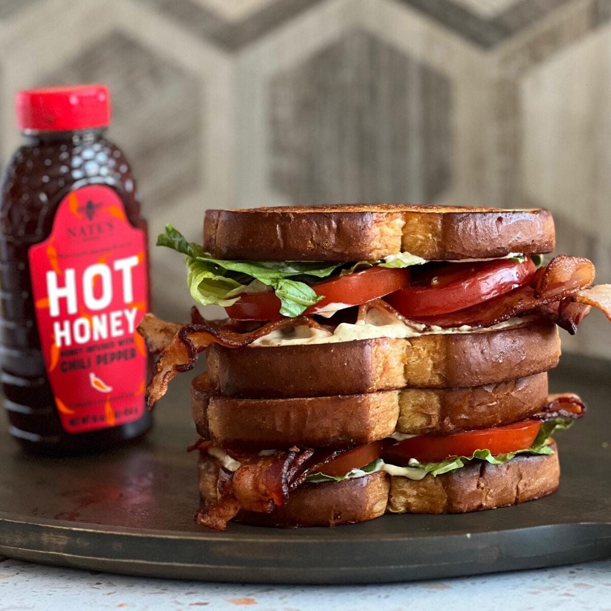A tower of BLT sandwiches with crispy bacon on a plate beside a bottle of Nate’s Hot Honey.