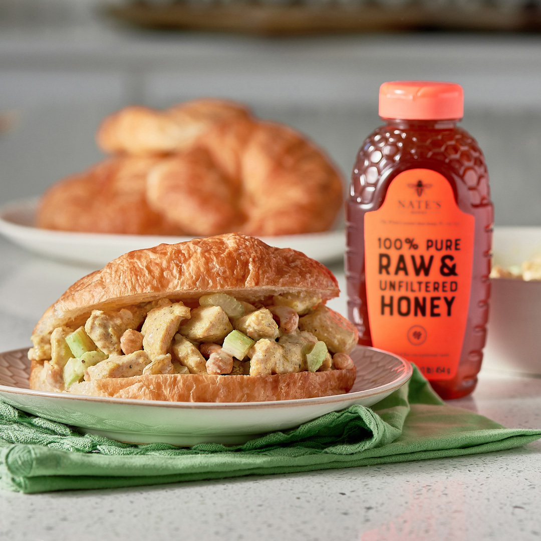 Curried chicken salad croissant sandwiches with a bottle of Nate’s Honey on a kitchen counter.