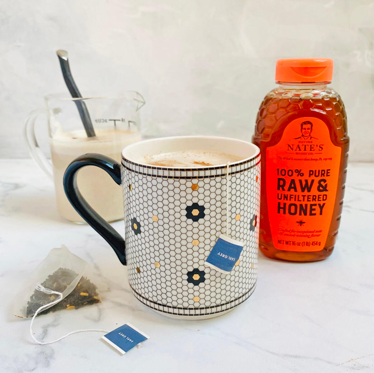 A mug with a floral pattern and a glass of milk next to a bottle of Nate’s honey, with tea bags on a marbled surface.