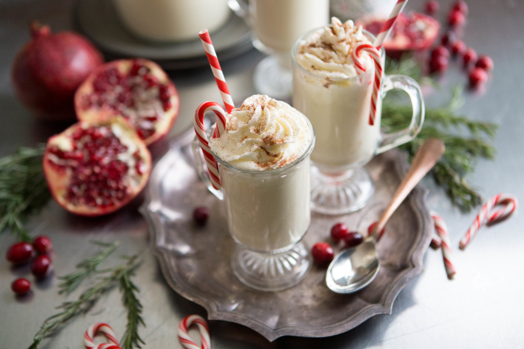 Eggnog in clear mugs with whipped cream, candy canes, festive straws, and fruit garnishes on a dark tray.
