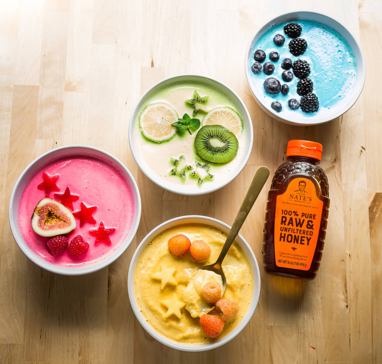 Colorful smoothie bowls with fruits and Nate's honey bottle on wooden surface.