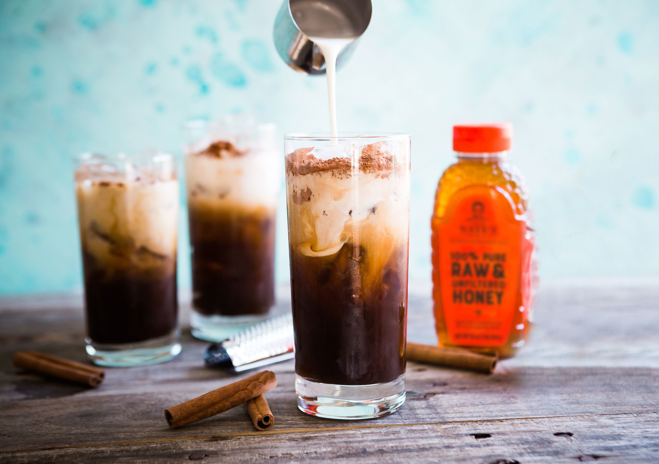 Pouring cream into iced coffee, with more glasses, cinnamon and a bottle of Nate's honey.