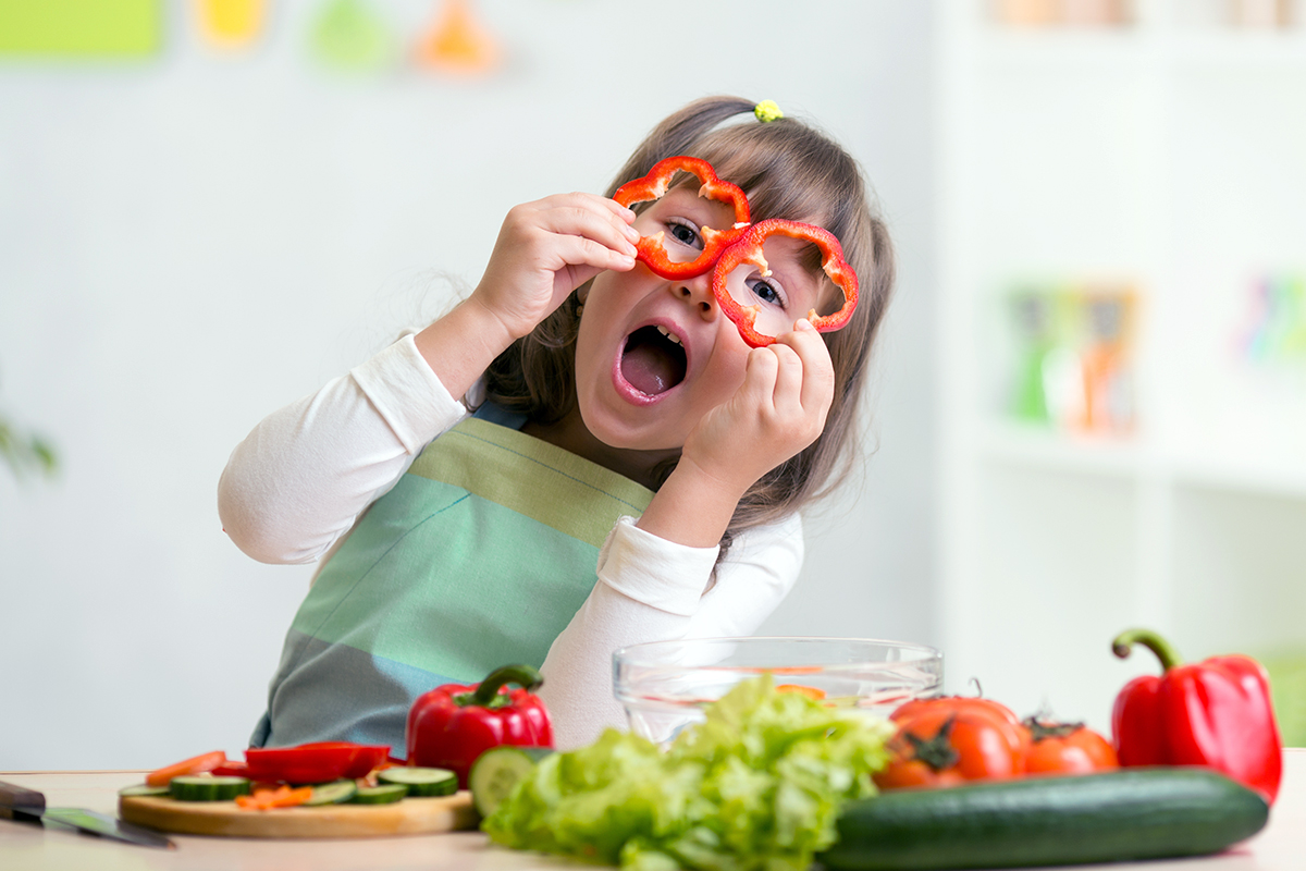 Child playing with red bell pepper slices as glasses, surrounded by fresh vegetables on a kitchen counter.