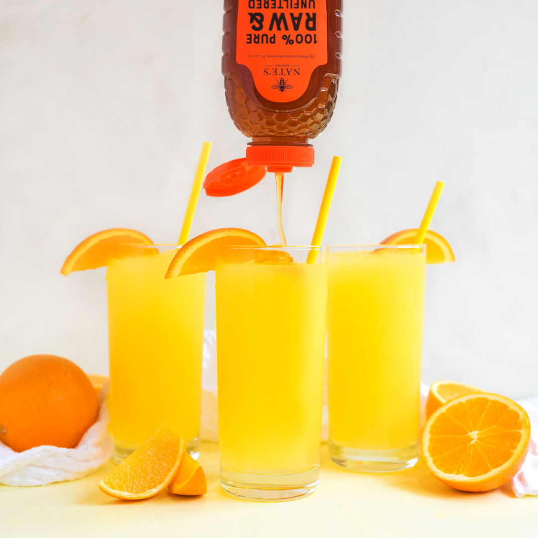 Three glasses of citrus slushie with straws and orange slices with Nate's Honey being drizzled into one.