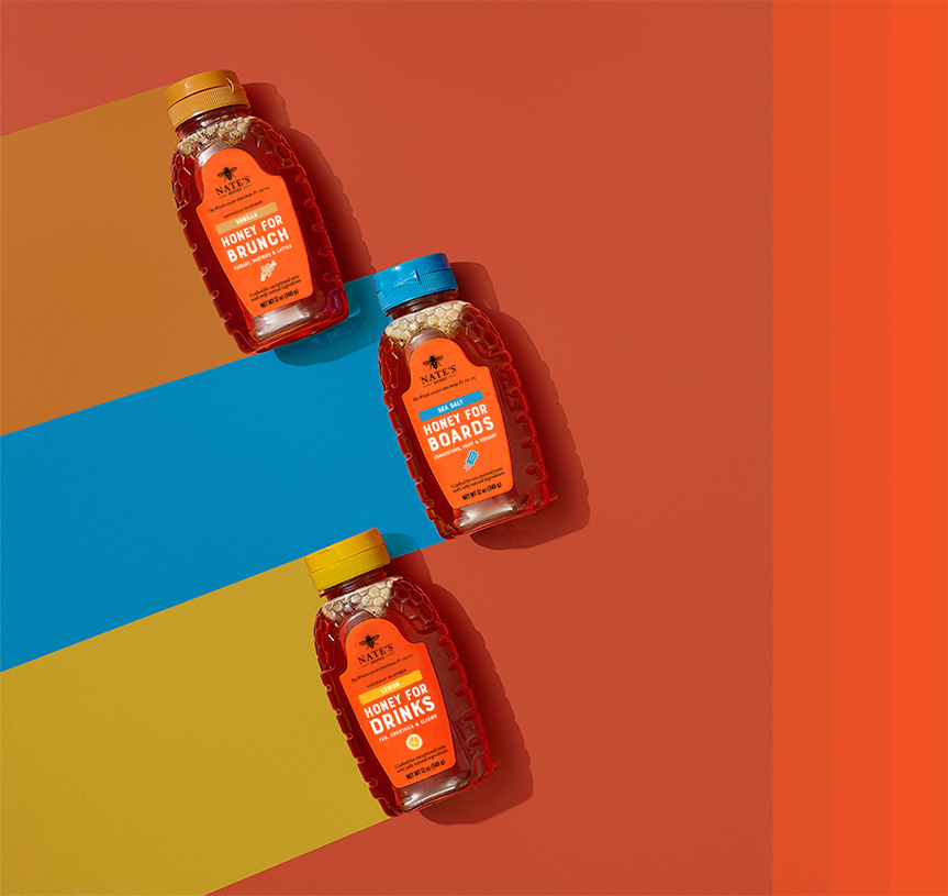 Three bottles of Nate’s Flavored Honey — Honey for Brunch, Honey for Boards and Honey for Drinks — arranged on a colorful striped background.