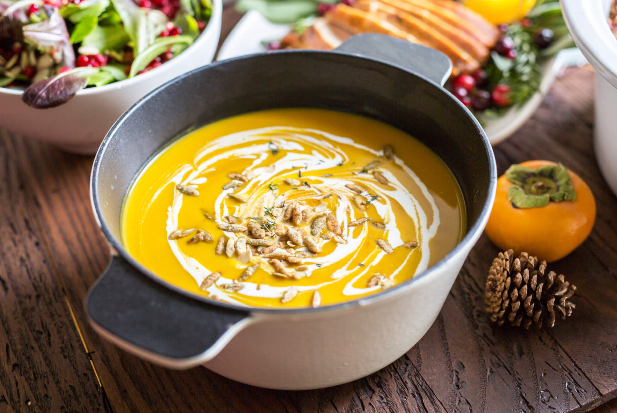 Pumpkin soup in a pot, cream swirl and seeds on a wood table with fall decor.