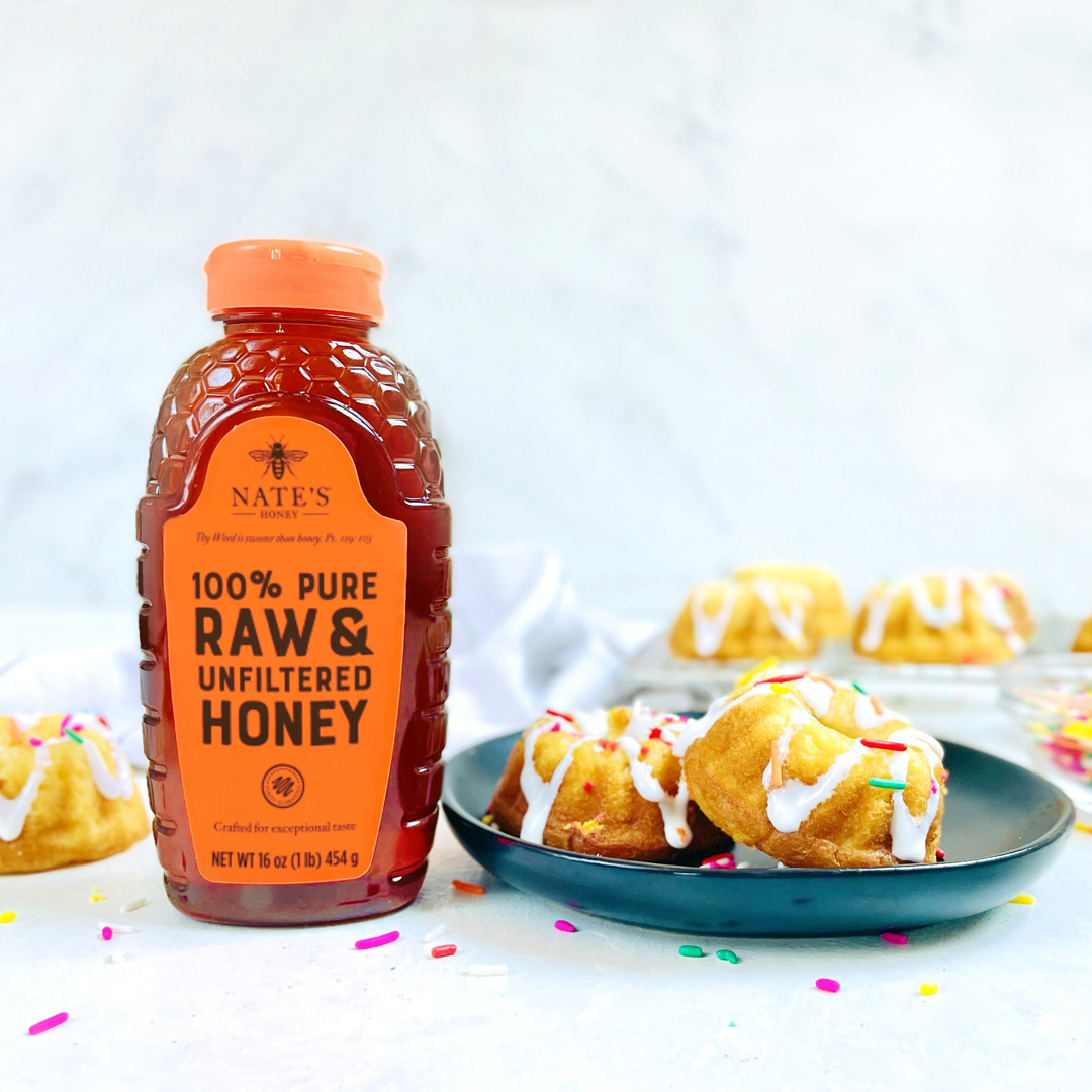 A bottle of Nate's Honey with a plate of frosted mini bundt cakes and colorful sprinkles on a marble surface.