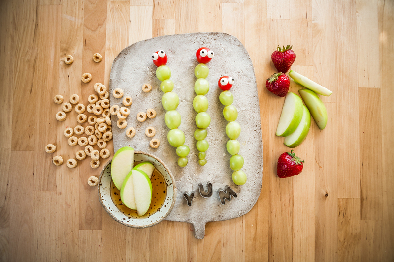 Food art on a cutting board spelling YUM, with grapes as caterpillars, cereal and apples with honey, on a wood surface.