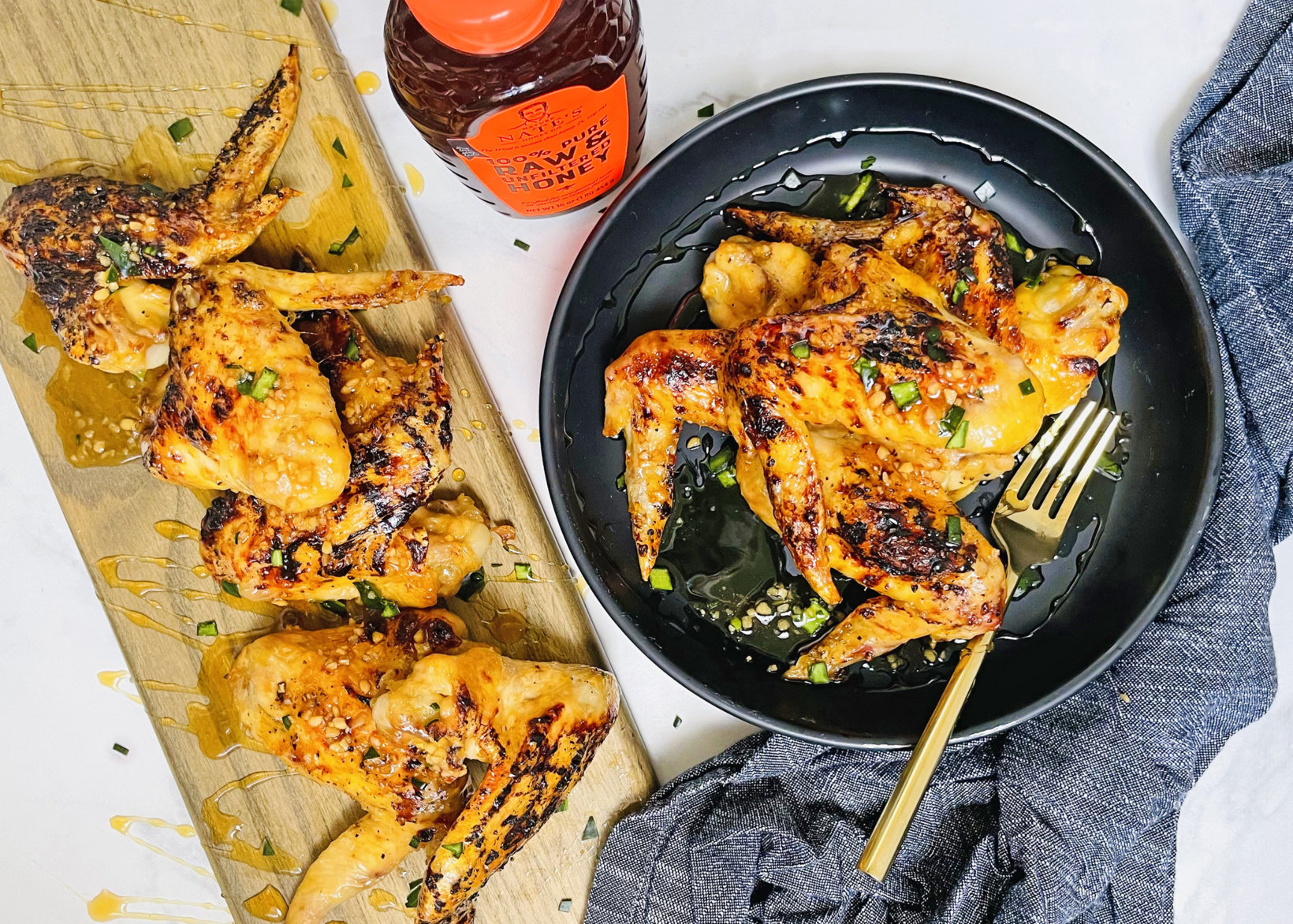 Honey garlic chicken wings on a plate and board, with a bottle of Nate’s honey and a gray cloth.