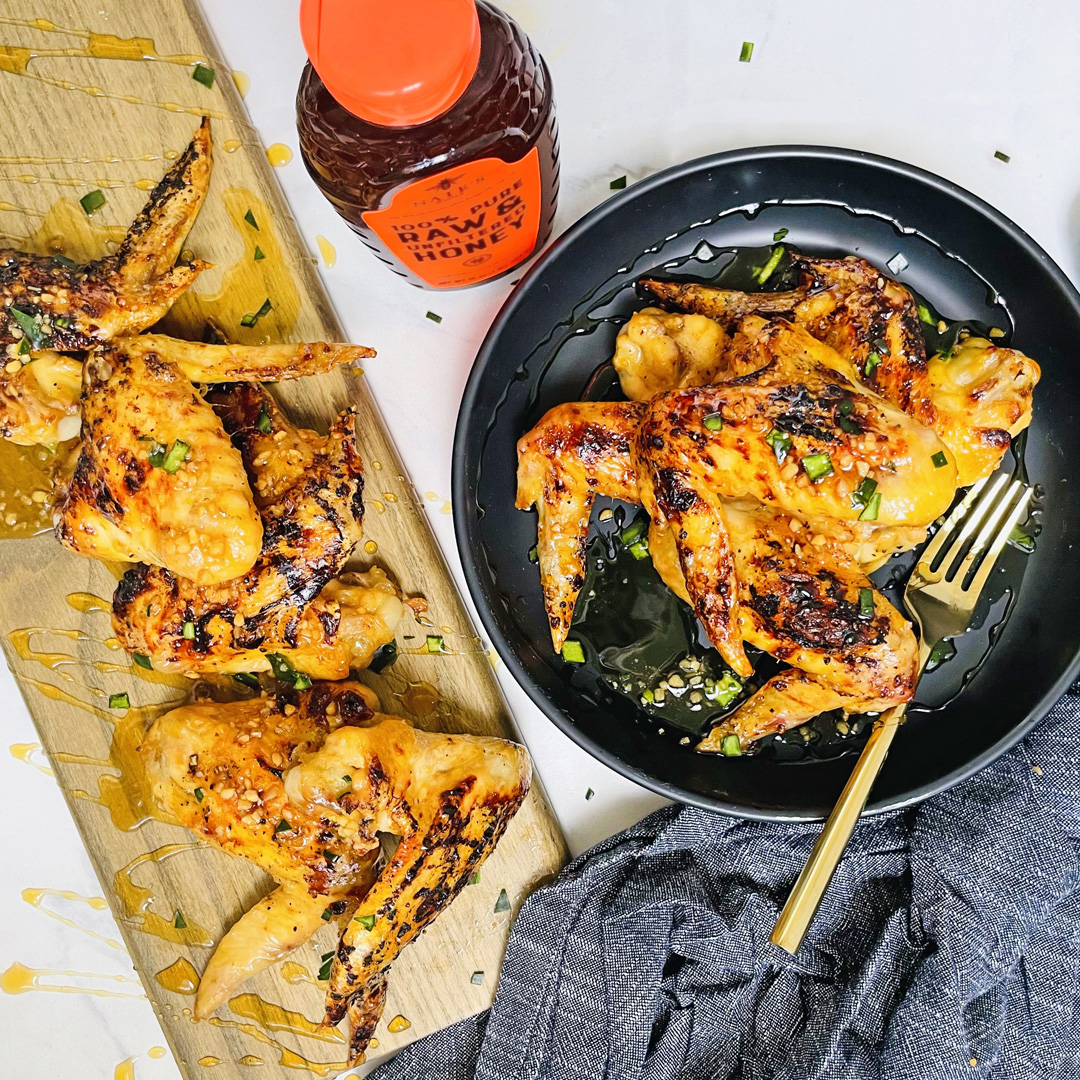 Honey garlic chicken wings on a plate and board, with a bottle of Nate’s Honey and a gray cloth.
