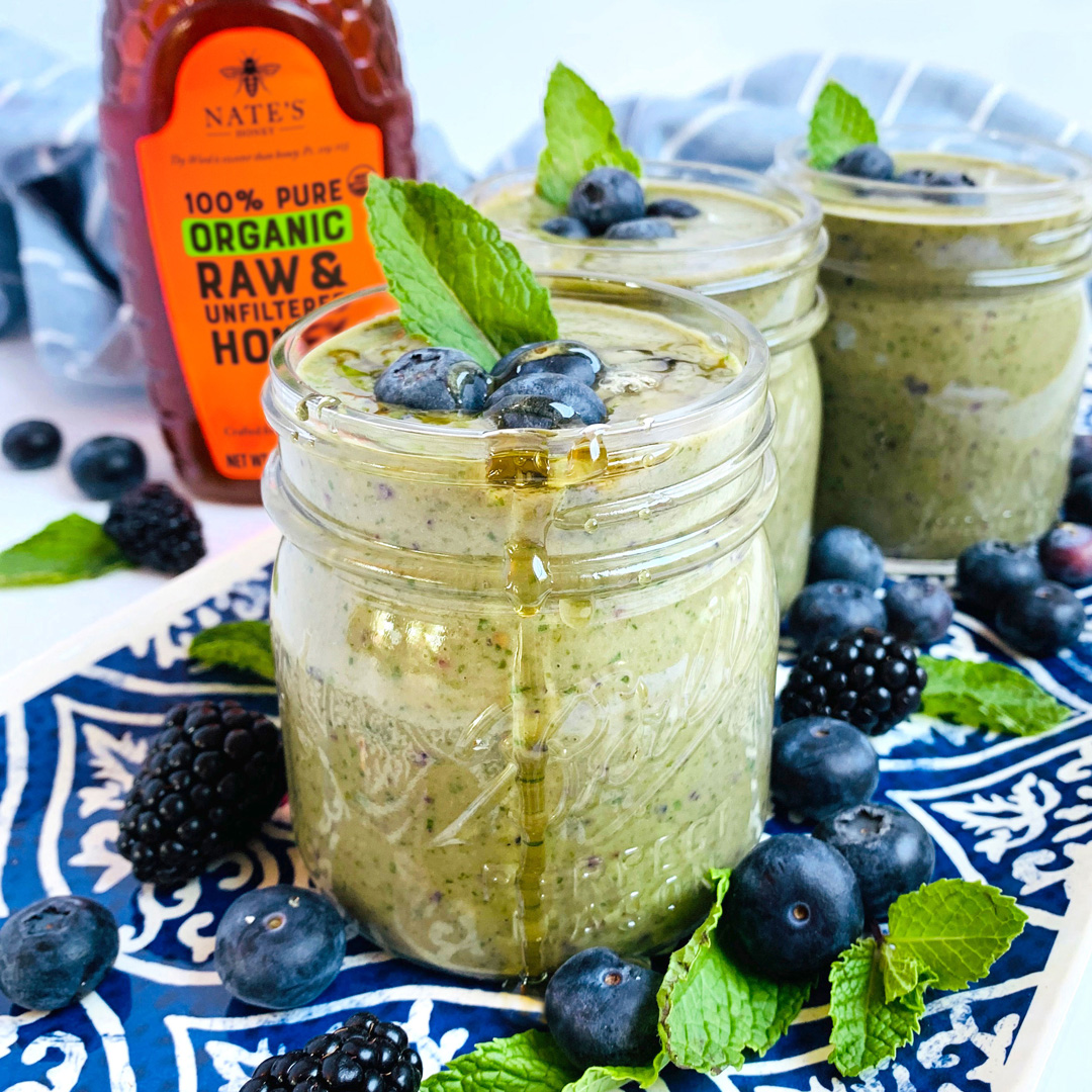 Green smoothie in jars, organic Nate's Honey bottle beside blueberries, blackberries and mint on blue patterned cloth.
