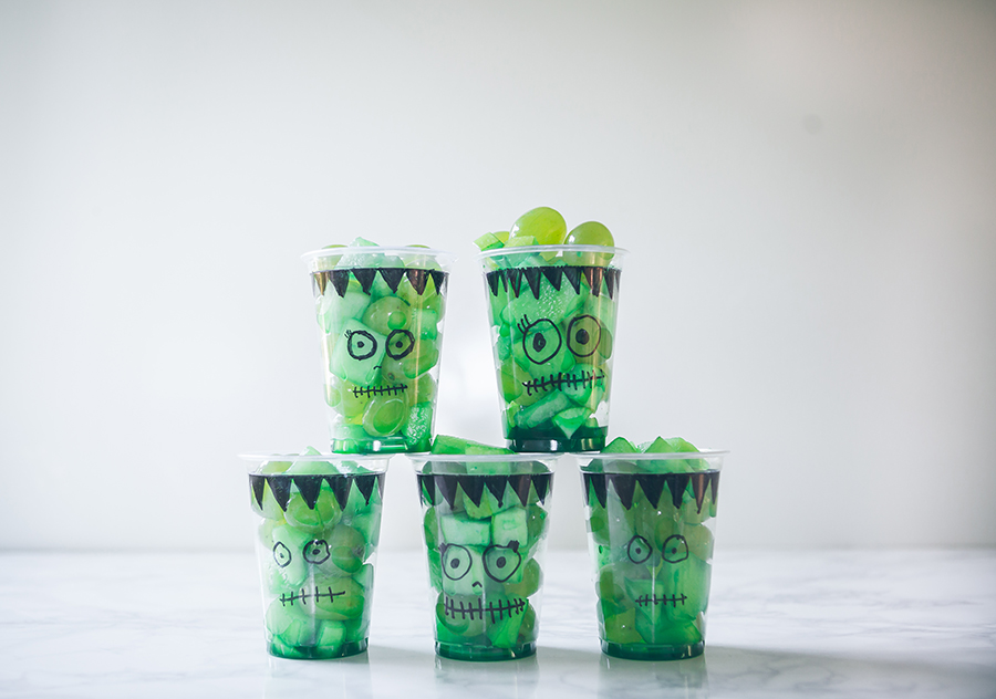 Green fruit cups stacked in a pyramid, decorated with Frankenstein faces for a fun Halloween treat.