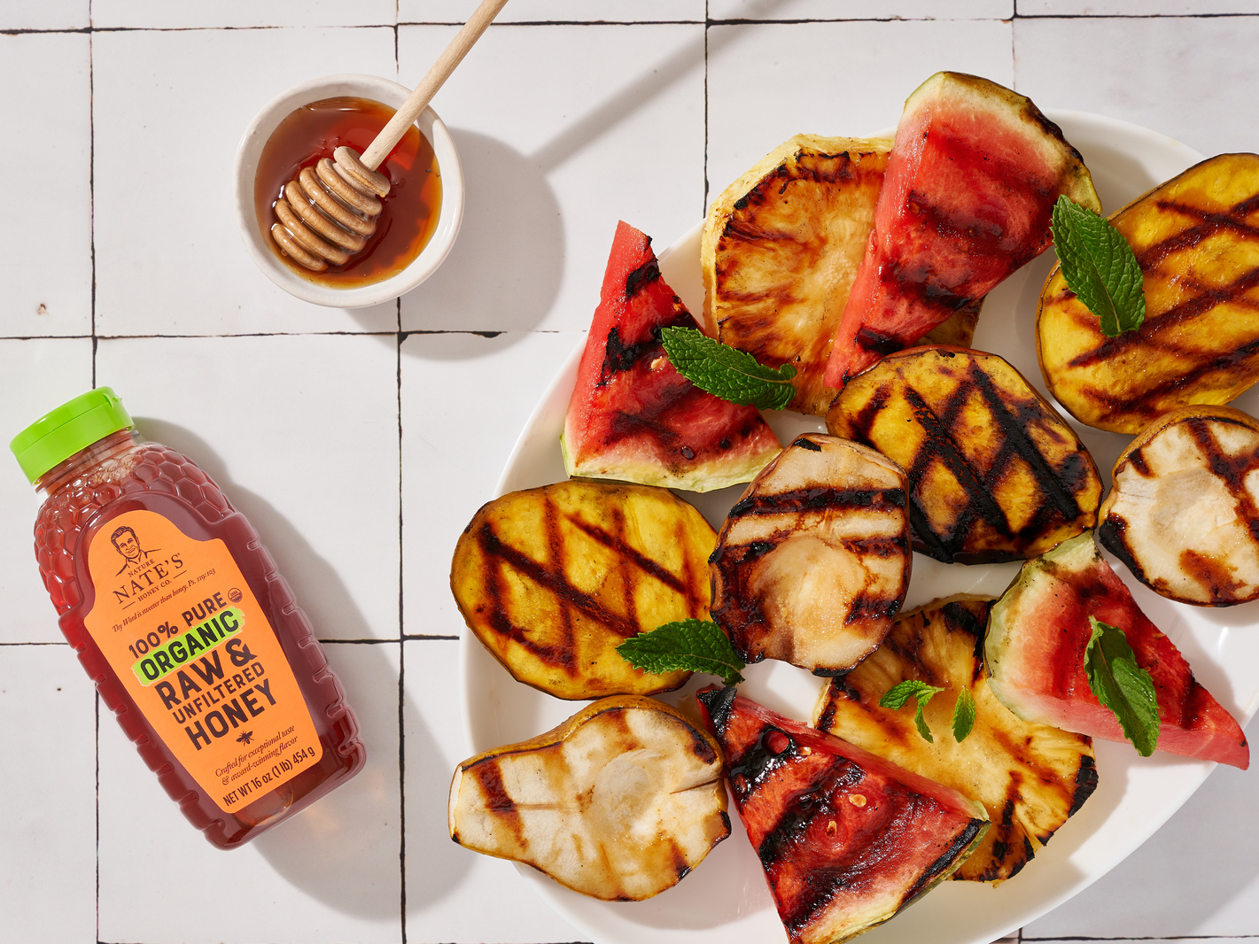 Grilled fruit slices on a plate with mint leaves, a honey dipper and a bottle of Nate’s organic honey.