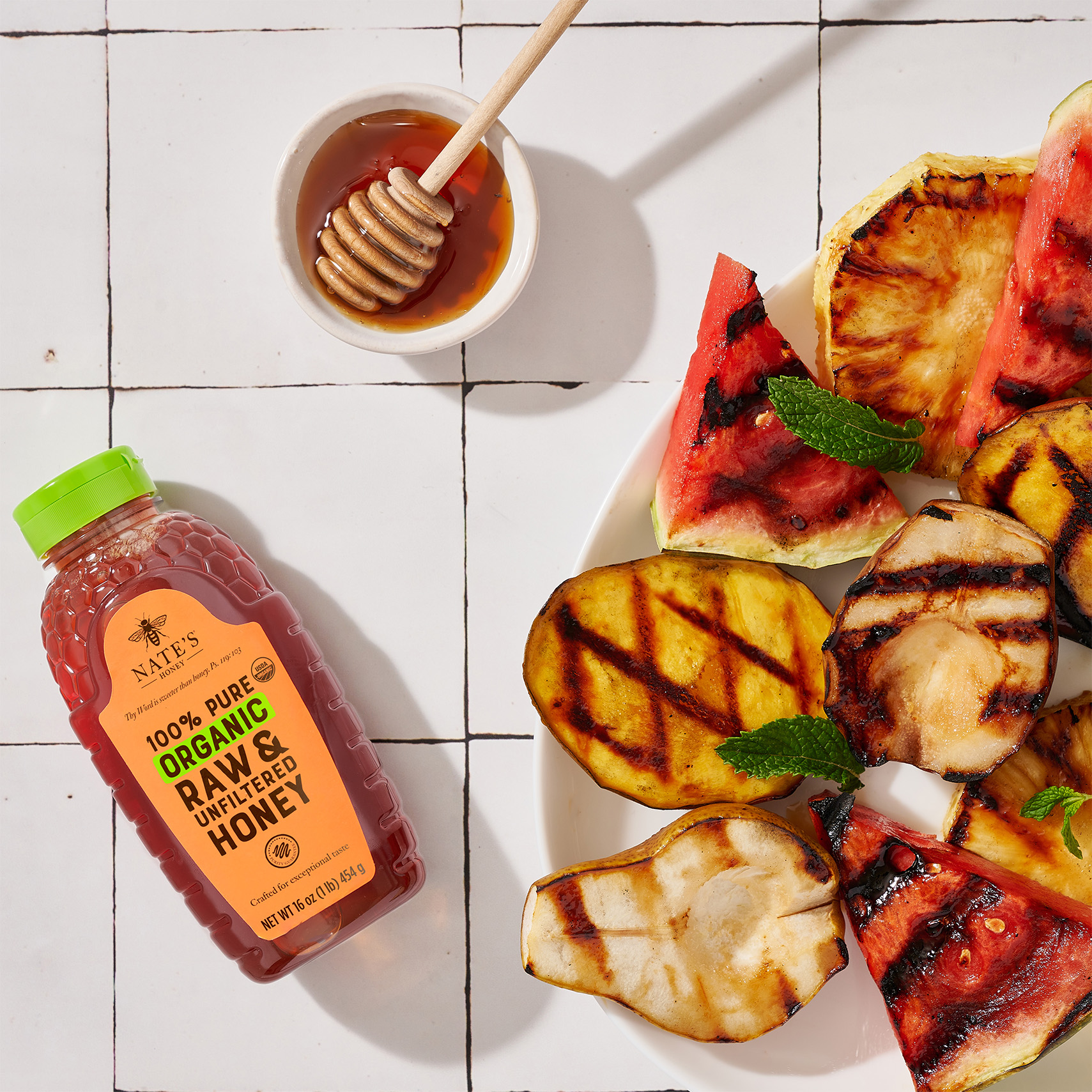Grilled fruit slices on a plate with mint leaves, a honey dipper and a bottle of Nate’s Organic Honey.