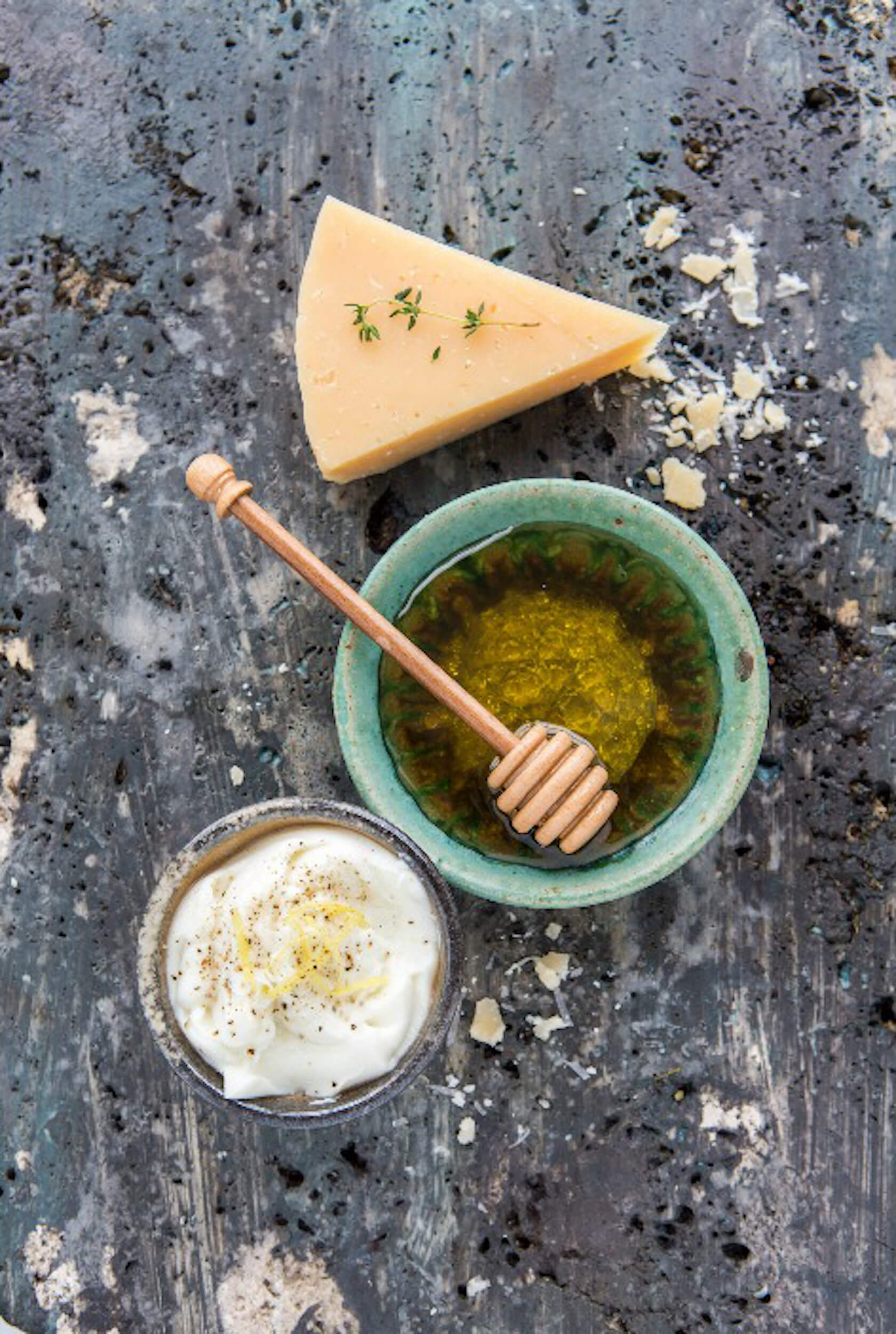Cheese wedge, honey in bowl with dipper, and soft cheese in a small bowl on a speckled backdrop.