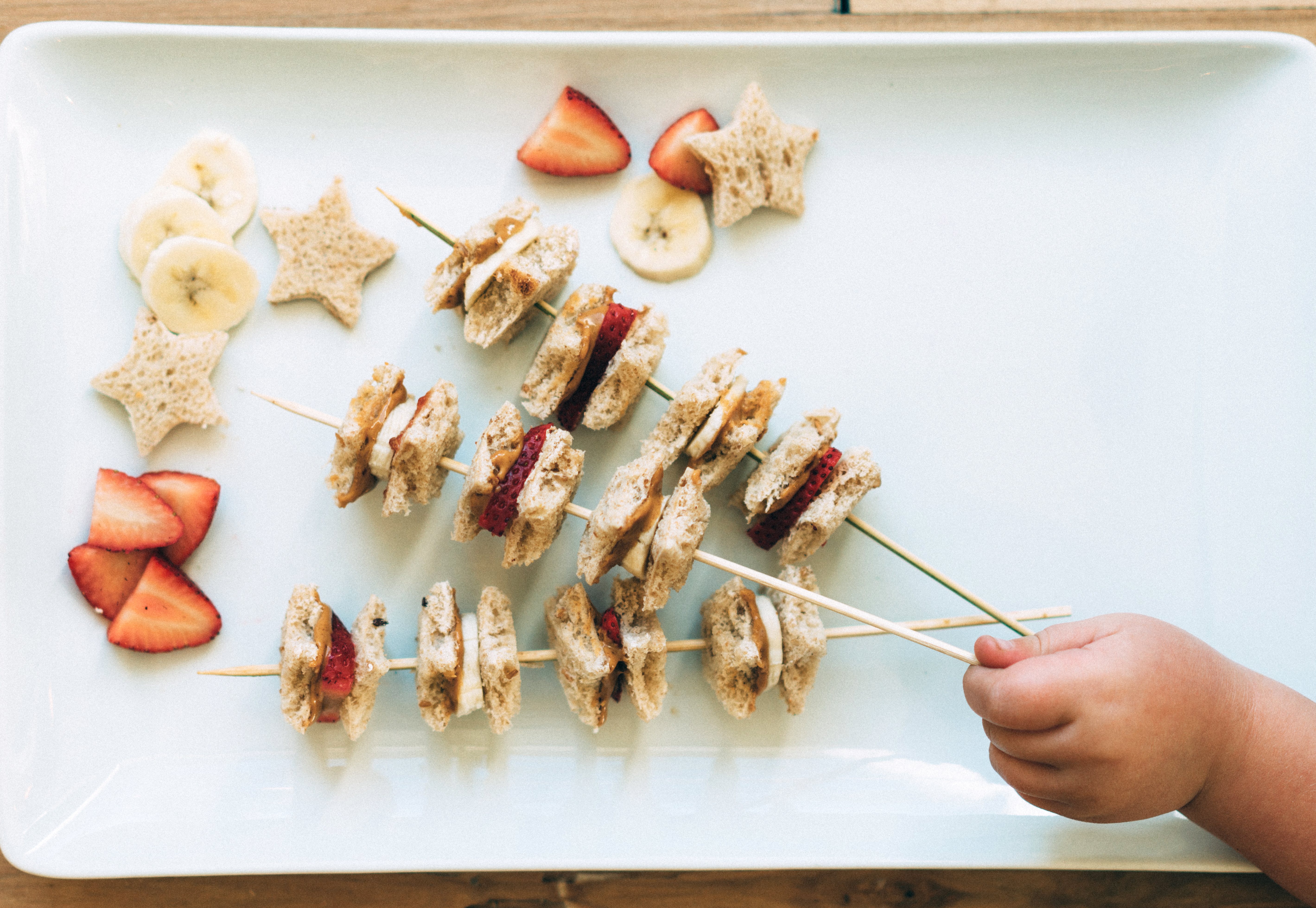 Child's hand picks skewer with banana, strawberry, sandwich bites, and star-shaped bread on white platter.