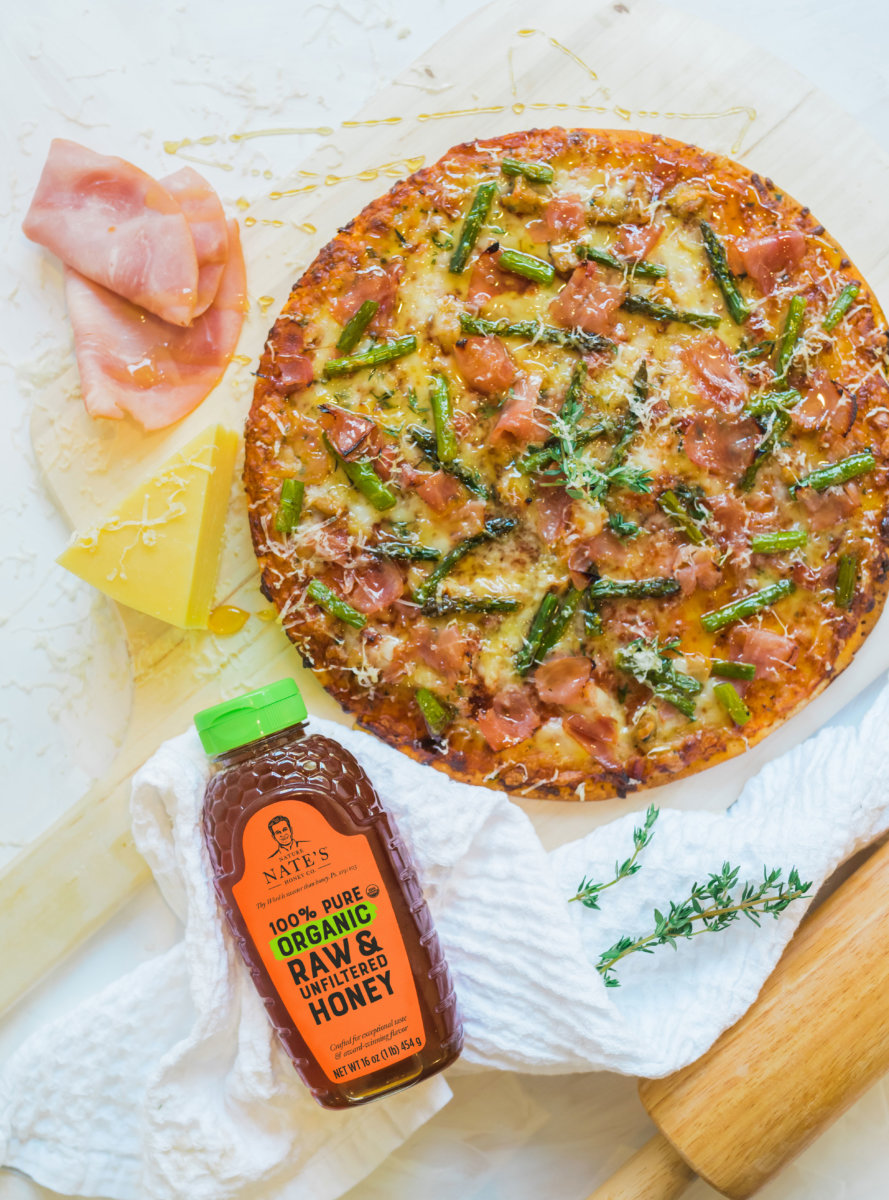 Asparagus and ham pizza with a bottle of Nate's organic honey and cheese on a white crinkled cloth.