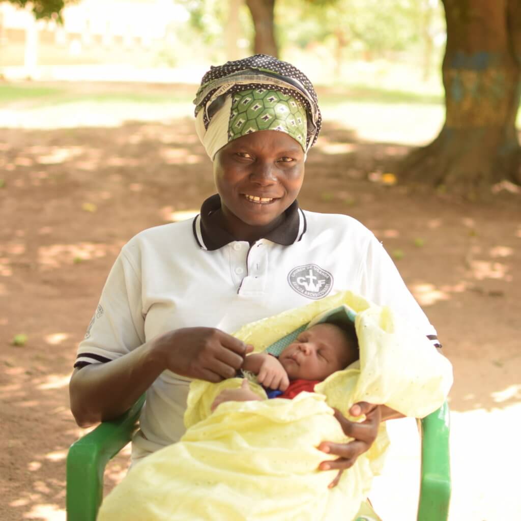 A woman in a headscarf smiling, holding a sleeping baby wrapped in a yellow blanket, seated outdoors.