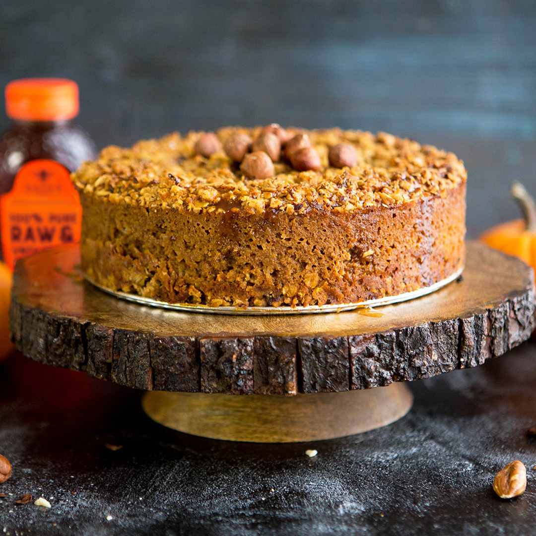 A nut-topped cake on a wood slice stand, with Nate's Honey and pumpkins, on a dark backdrop.