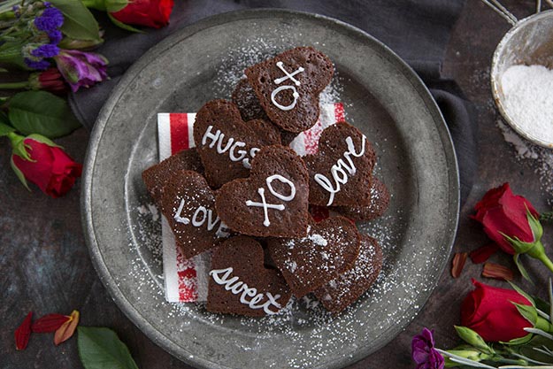 Heart-shaped chocolate brownies with romantic words written with icing on top, surrounded by roses.