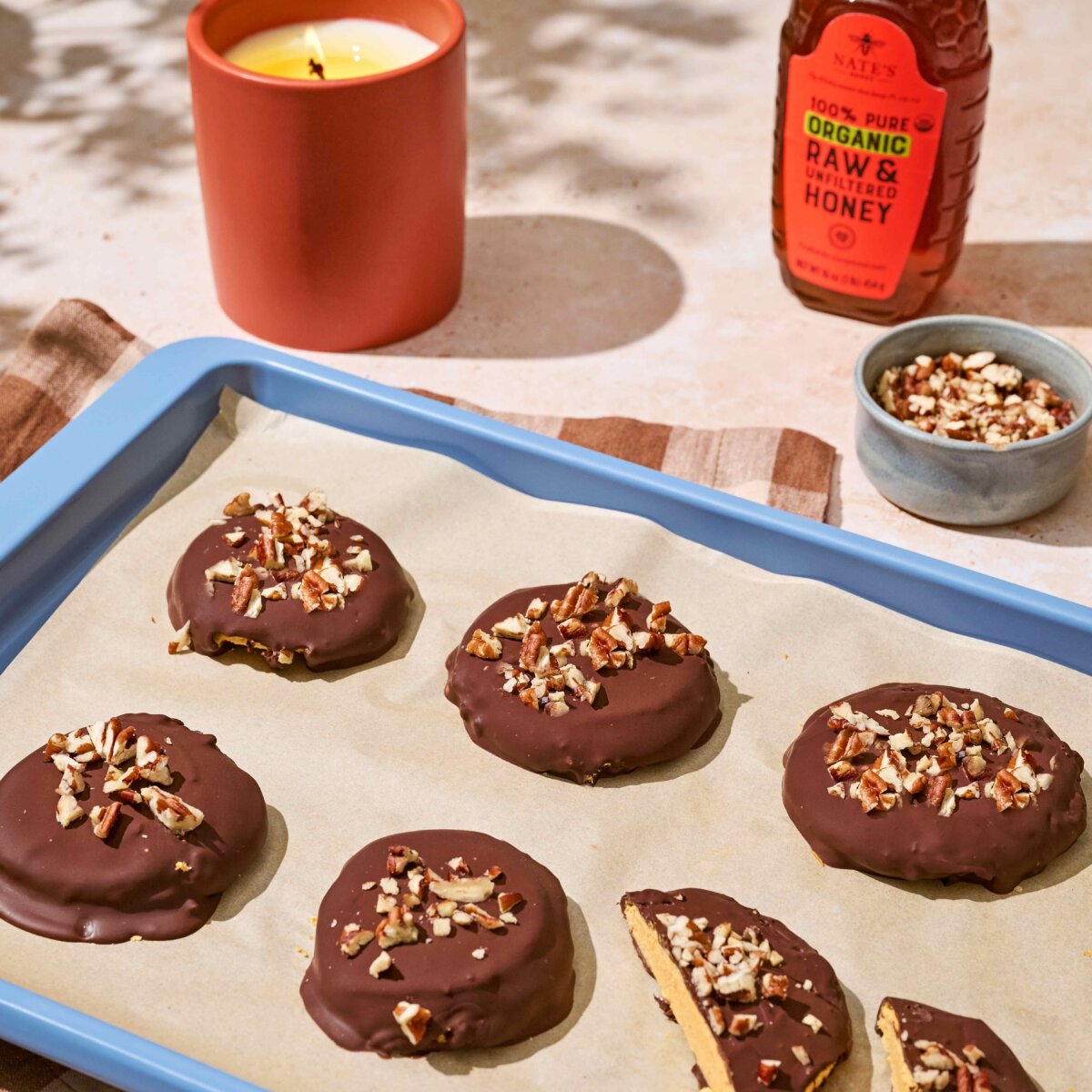 Sheet pan holding chocolate‑coated round dessert topped with chopped nuts, with a bowl of nuts and a bottle of honey in the background.