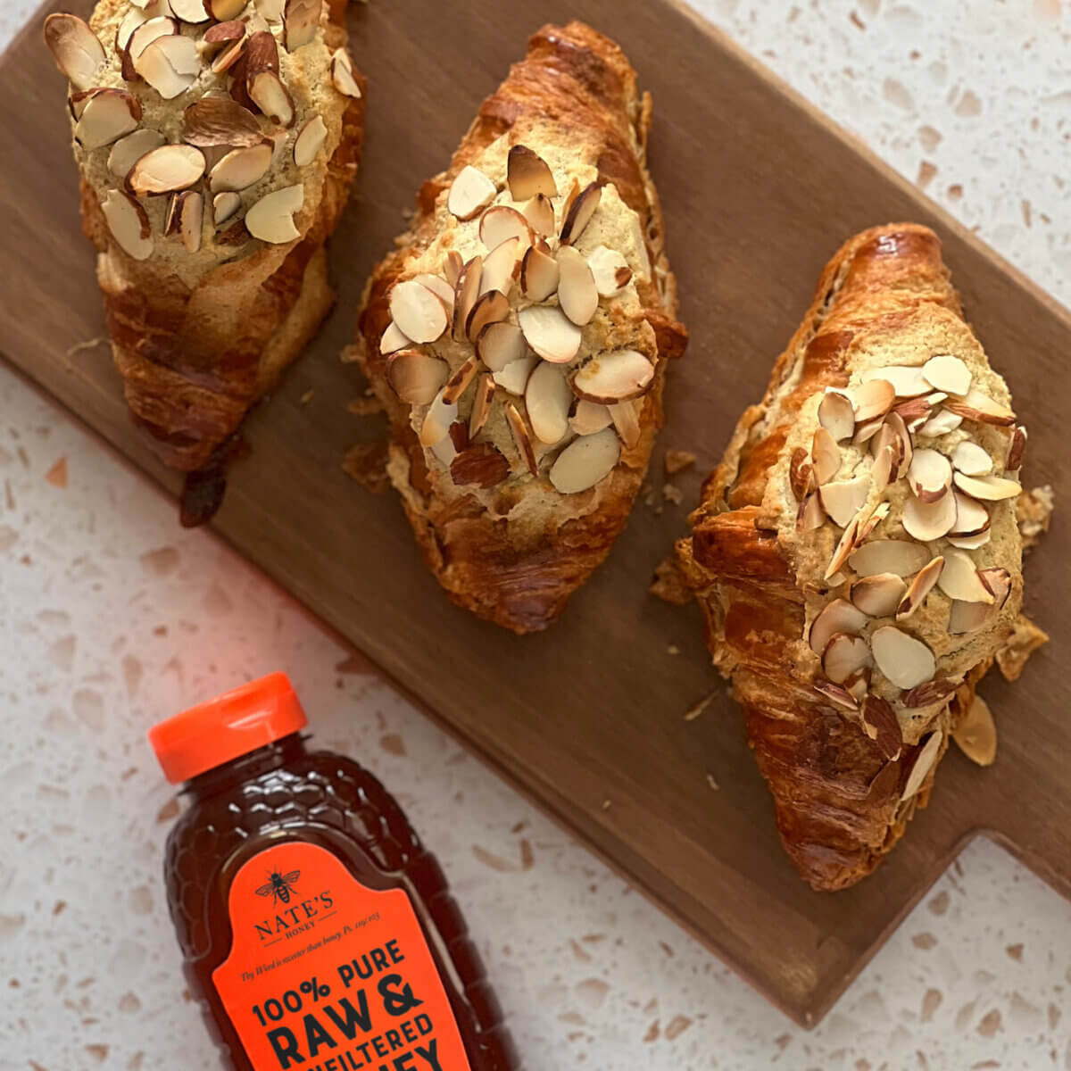 Three honey almond croissants on a board next to a Nate’s honey bottle.