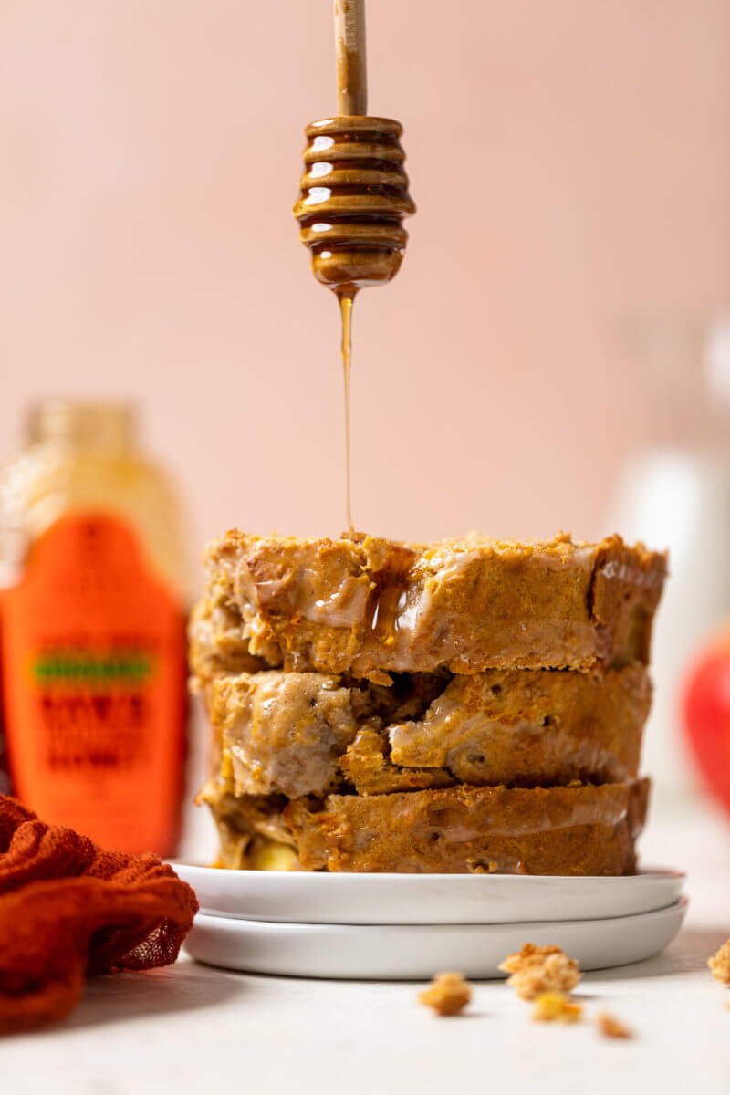 A stack of apple carrot bread slices with dripping honey next to  Nate’s organic honey bottle on a pink background.