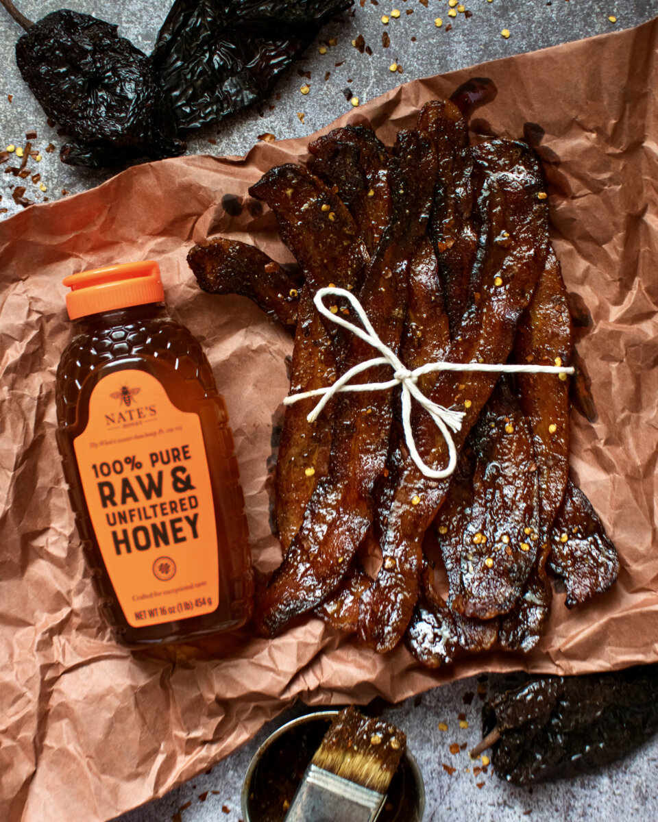 Honey-glazed bacon and a bottle of Nate's Honey with an orange cap on a speckled counter.