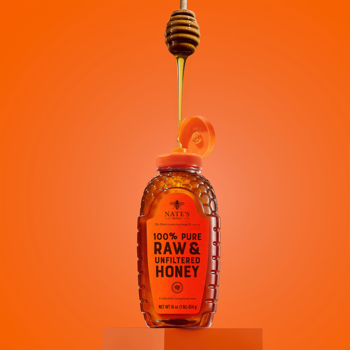 Bottle of honey sitting on orange block with honey dipper drizzling over it.