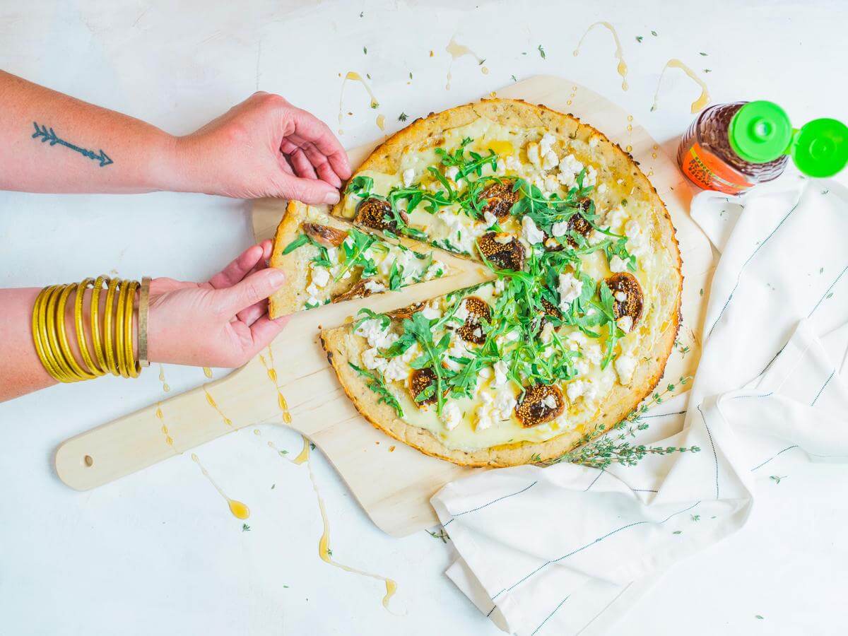 A hand taking a piece of honey, fig and mascarpone flatbread, beside a bottle of Nate’s organic honey and a napkin.