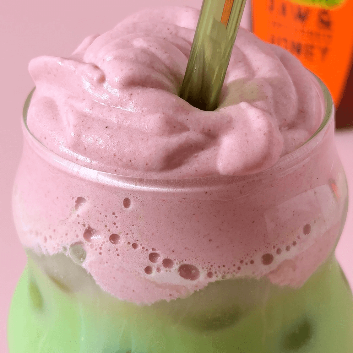 Glass with a green matcha latte and strawberry cold foam on top. Green straw in the latte with a Nate’s organic honey bottle in background.