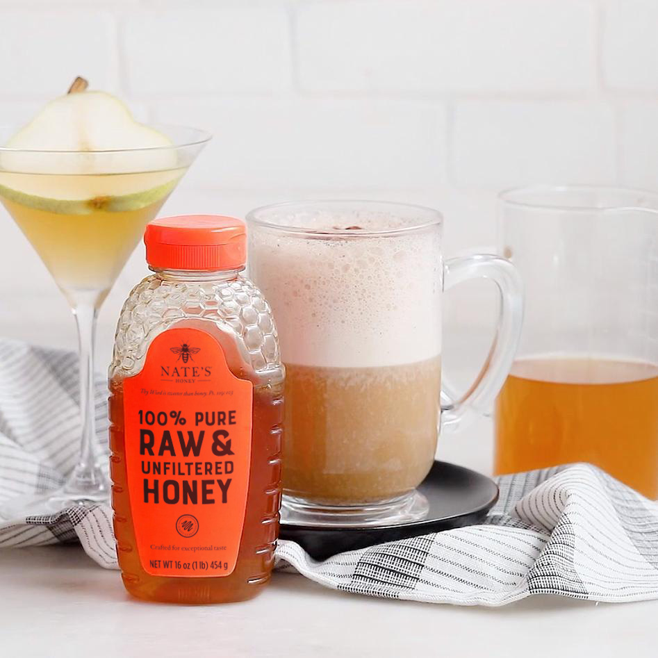 A frothy coffee latte in a mug with a bottle of Nate’s Honey, a cocktail and a checkered cloth backdrop.